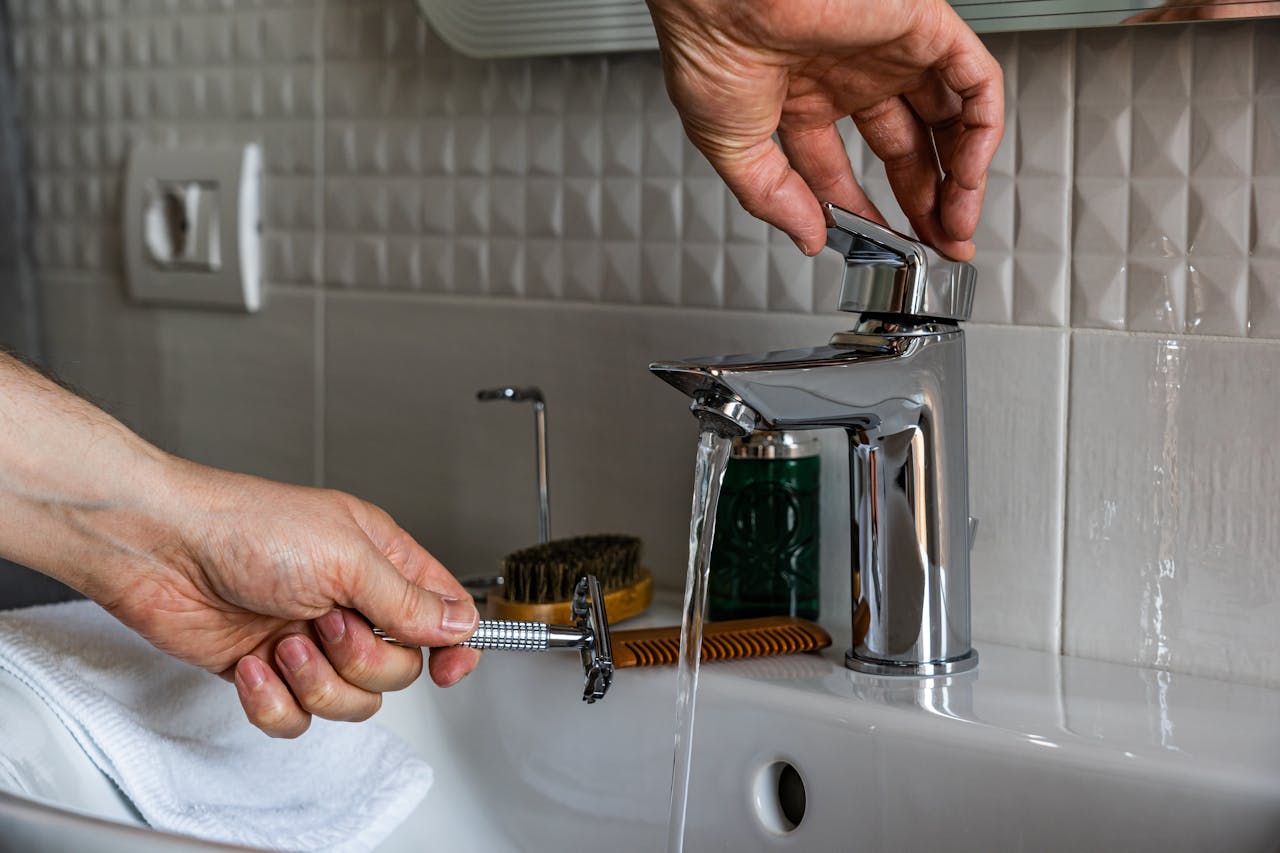 A close-up of hands using a faucet to rinse a razor in a modern bathroom sink.